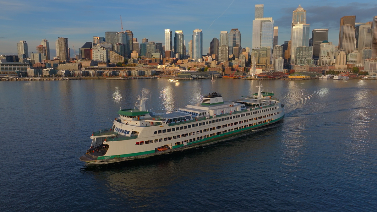 A large ferry sails across calm water approaching a city skyline with tall skyscrapers under a clear blue sky at sunset.
