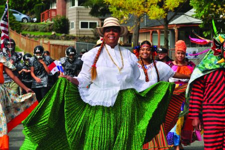 A group of people participating in a colorful parade. A woman in the foreground wears a vibrant green skirt, white lace top, and a straw hat, smiling while holding up her skirt. Others in colorful costumes and masks are visible, with a backdrop of trees and houses.