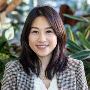 A person with shoulder-length dark hair smiles at the camera, standing in front of green plants. They are wearing a plaid blazer over a light top.