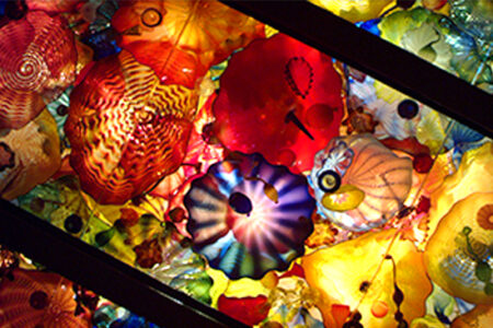 A vibrant ceiling display of colorful, intricately designed glass art pieces. The glass is sculpted into organic shapes with swirling patterns in shades of red, blue, orange, yellow, and purple, creating a kaleidoscopic effect when illuminated.