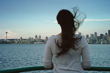 A person with long hair stands on a boat, looking at the Seattle skyline across the water. The Space Needle is visible in the distance, and the sky is clear with a few clouds.