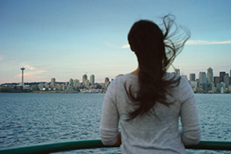A woman with long hair stands on a boat, looking at a city skyline across the water. The wind blows her hair, and a prominent tower is visible in the distance against a clear sky.