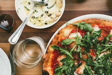 A pizza topped with arugula sits on a wooden table next to a bowl of pasta with a spoon. A glass is nearby, and a pair of sunglasses is placed beside the dishes.