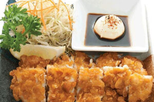 A plate of breaded and fried cutlet slices, served with shredded cabbage, carrot, parsley, a lemon wedge, and a small dish of dipping sauce topped with a dollop of mayonnaise and chili flakes.
