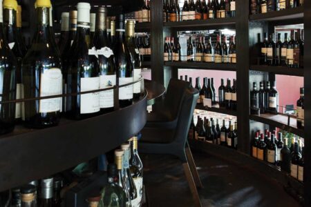 A curved wine cellar with multiple shelves filled with a variety of wine bottles. The dimly lit setting features a small chair tucked in the corner, creating a cozy ambiance. The floor is made of dark concrete, and the shelves are predominantly wooden.