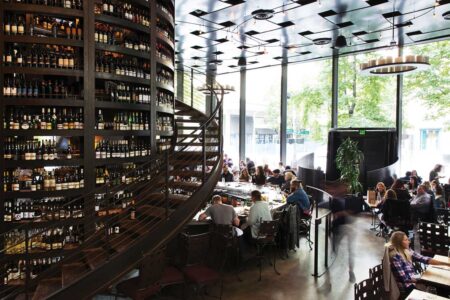 A modern, bustling restaurant interior with floor-to-ceiling windows. Large circular shelves filled with wine bottles surround a spiral staircase. Diners sit at tables, enjoying meals beneath unique, contemporary lighting fixtures.