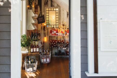 A cozy wine shop features shelves with bottles of wine, including a variety labeled "Rosé Vibes." Baskets sit on the wooden floor with more bottles. A mirror and small plants add to the inviting interior. Sunlight casts gentle shadows.