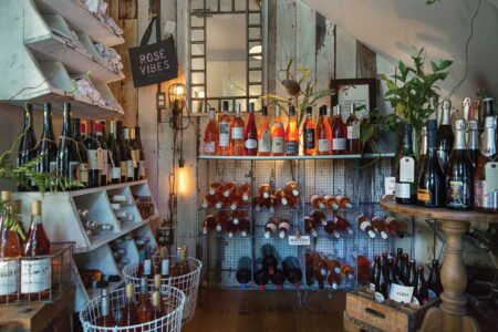A cozy wine shop with rustic decor features various bottles of wine displayed on wooden shelves and wire racks. A mirror and hanging light add to the ambiance, with a 
