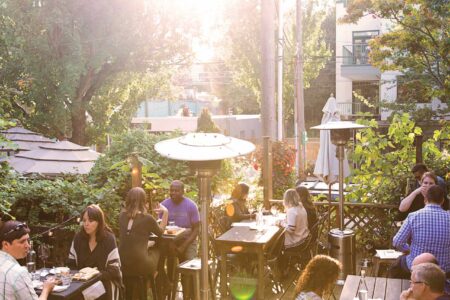 A lively outdoor restaurant patio with people dining at wooden tables. Tall patio heaters are scattered around. Sunlight filters through trees, creating a warm, inviting atmosphere. Bushes and greenery surround the deck, enhancing the cozy setting.