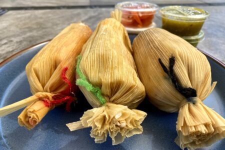Three steamed tamales aligned on a plate with jars of condiments in the background.