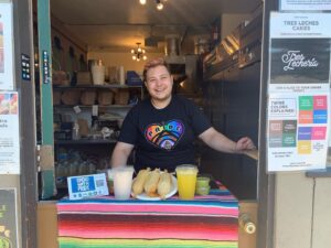 A person in a black t-shirt stands behind a counter draped in a colorful blanket. Two drinks and a plate of tamales are placed on top.