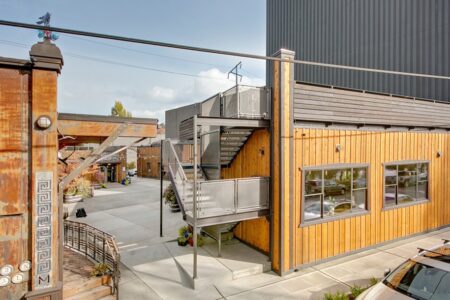 An industrial building with a wooden exterior, metal staircases, and large windows. The surrounding area includes concrete ground, railings, and a parked car. The sky in the background is cloudy.