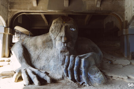 A large stone sculpture of a troll with a single eye and a rugged appearance is situated under a concrete bridge. The troll's hand holds a car, and its expression is both whimsical and imposing. Dirt and sparse foliage surround the sculpture.