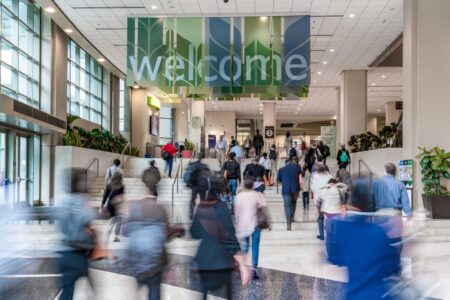 A bustling indoor scene shows people walking up a flight of stairs toward a large sign that reads 