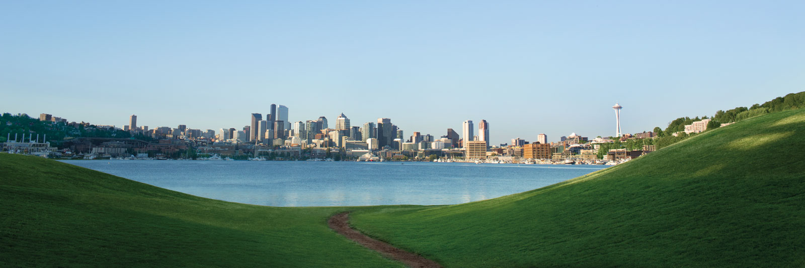 A panoramic view of a city skyline across a vast body of water, framed by gently sloping green hills. The city features numerous skyscrapers and a prominent tower with a needle-like top.