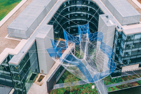 Aerial view of a modern, L-shaped building with a large sculpture resembling a spider web and hand in the courtyard. The building features glass walls and green rooftops, surrounded by manicured lawns.