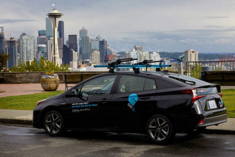 A black sedan parked in front of Kerry Park in Seattle with the city skyline in the background. The car has bright blue details including the GIG Car Share logo and a rack on top of the car. A white and blue decal on the driver's door reads, "Late for yoga class? Get there with GIG Car Share. Drive 1 hour free. use promo 'GIGTRIP'".