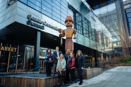A group of men and women stand in front of a tall wooden statue. A steel and glass building is in the background.