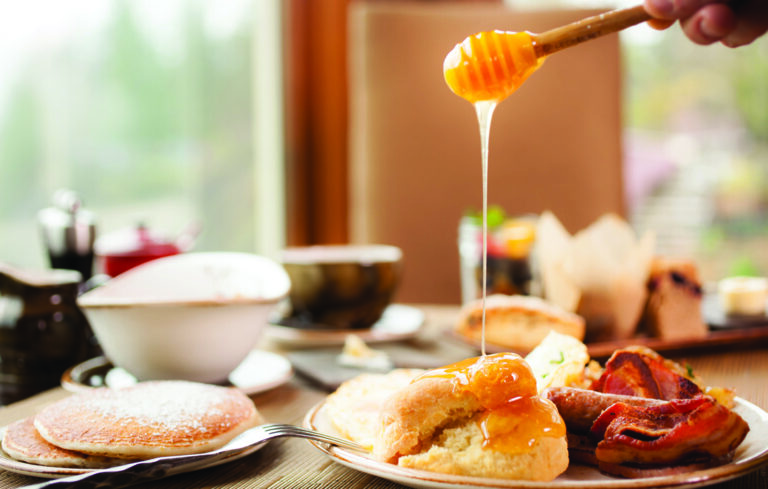 A breakfast spread with honey being drizzled over a biscuit. The table includes pancakes, bacon, a bowl, and assorted pastries, set near a window with a blurred view of greenery outside.
