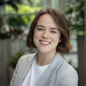 A person with short brown hair smiles while wearing a gray blazer and white shirt. They are indoors with greenery and a window in the background.