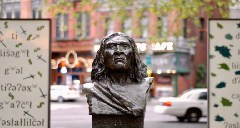 A bronze bust of Chief Seattle is centered, surrounded by decorative panels with indigenous motifs and greenery. The background features a blurred urban scene with visible cars and building lights.