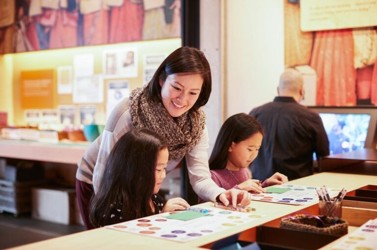 A woman smiles while helping two young girls with an activity at a table in a brightly lit, interactive museum—one of the top things to do in Seattle. Another person works at a computer in the background.