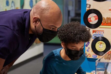 A man and a child, both wearing masks, closely observe a museum exhibit featuring retro diner decor and vinyl records. The child, with curly hair, seems particularly curious.