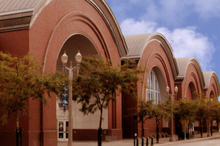 A row of large, historic brick arches with framed glass windows against a blue sky. Trees and vintage-style street lamps line the sidewalk in front of the building.