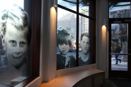 Black and white photos of children are displayed on large window panels inside a building. Through the windows, an urban city scene with pedestrians and tall buildings is visible. The interior has a curved bench beneath the windows.