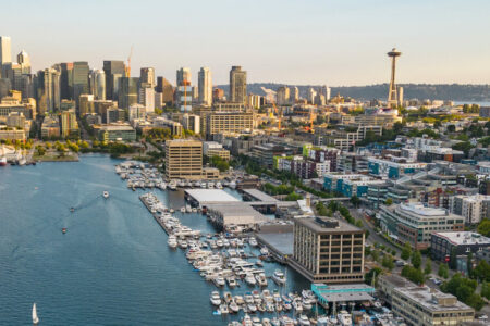 Aerial view of a city skyline with numerous skyscrapers and a prominent tower. A large body of water with docks and boats is in the foreground. The landscape is lush and green, with hills in the background.