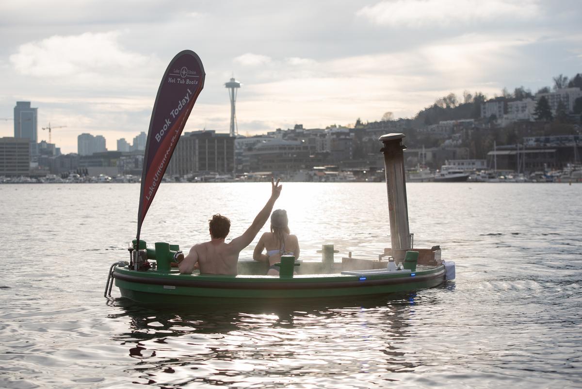 Two people relax in a round hot tub boat on a lake at sunset, with city buildings and the Space Needle visible in the background. One person waves toward shore.