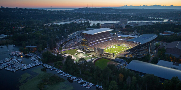 Aerial view of a Seattle sports stadium filled with spectators during an evening game. The stadium is illuminated, surrounded by water, trees, and a cityscape under a colorful sunset sky. Boats are docked nearby.
