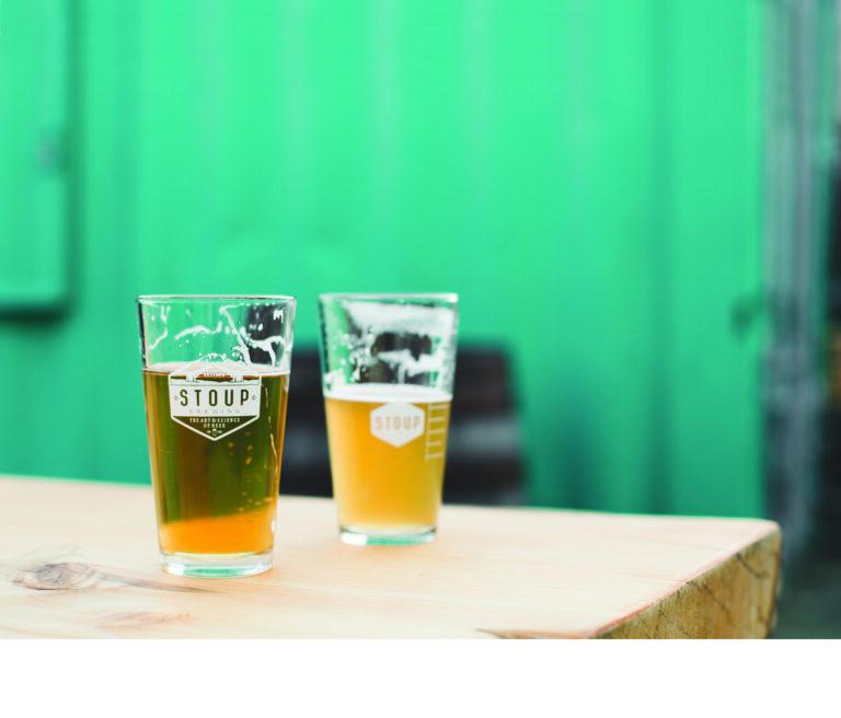 Two glasses of beer rest on a wooden table against a vibrant green background. Each glass has a logo and contains a different type of beer; one is a pale amber color, and the other appears slightly hazy and lighter.