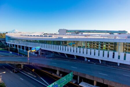 Exterior view of a large airport terminal with a modern design, featuring a long, curved building with many windows. Multiple highways and signs are visible in the foreground, and a clear blue sky is in the background.