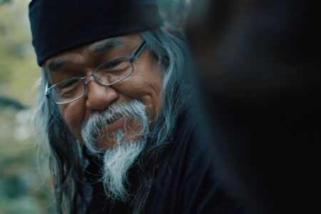 An elderly man with long gray hair and a beard, wearing glasses and a black bandana, smiles gently. The background is blurred, featuring greenery.