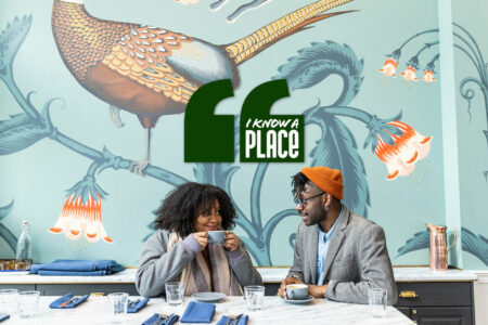 A couple enjoying coffee at a marble table in a cafe. The wall behind them is decorated with a large bird and floral design. A green sign with the text "I know a place" is displayed above them.