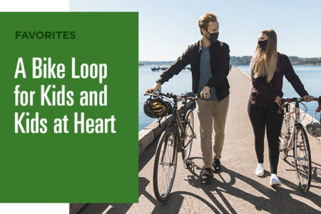 Two people wearing masks walk their bikes along a waterfront path. The image features the text: 
