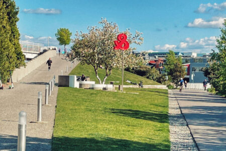 A park scene with a grassy hill and a pathway on each side. A sculpture of red roses stands near a blossoming tree under a blue sky with scattered clouds. People are walking and sitting, enjoying the sunny day.
