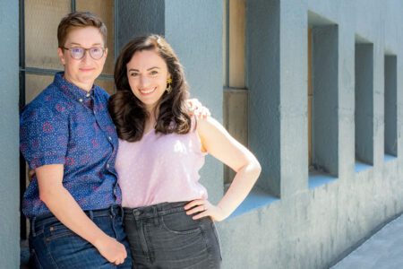 Two people stand closely together, smiling at the camera. The person on the left wears a blue patterned shirt and glasses, while the person on the right wears a light pink sleeveless top. They are leaning against a gray wall with square windows.
