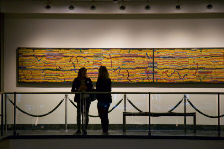 Two people stand in silhouette in front of a large, colorful abstract painting in a gallery. The artwork features vibrant horizontal lines and patterns. The gallery is dimly lit, highlighting the artwork.