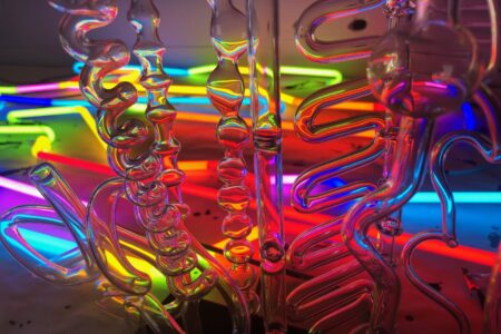 A vibrant display of colorful neon lights reflects through intricate glass sculptures. The lights glow in shades of red, orange, yellow, green, blue, and purple, creating a mesmerizing and abstract visual effect.