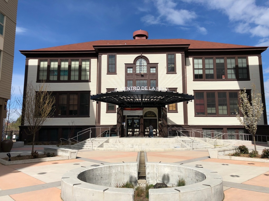 A large, two-story building with a red roof and cream-colored walls features a sign reading "CENTRO DE LA RAZA" above the entrance. The foreground includes a circular concrete structure and steps leading to the front door.