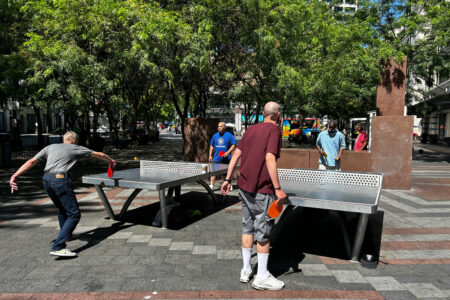 Four people play outdoor ping pong on two metal tables in a park surrounded by trees and buildings.