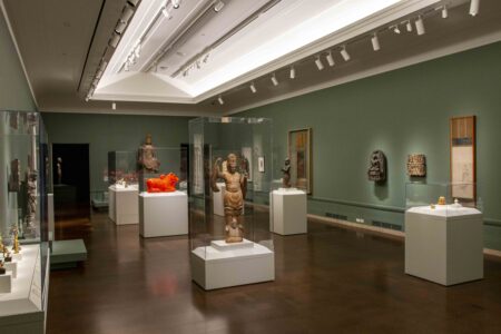 A museum gallery with green walls displaying various sculptures and artifacts in glass cases. The room is well-lit with ceiling lights, highlighting the artworks. The floor is dark and reflective, complementing the serene atmosphere.
