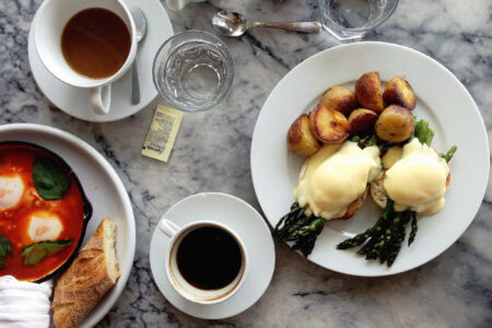 Top-down view of a breakfast table with plates of eggs Benedict with roasted potatoes, shakshuka with bread, two cups of coffee, a glass of water, and a small jar of condiments on a marble surface.