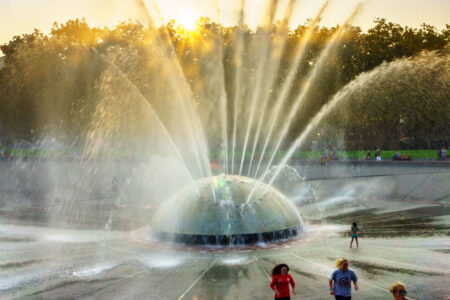 People walking in front of a large, spherical fountain with multiple water jets spraying upwards. The sun is setting behind trees in the background, casting a warm glow over the scene.