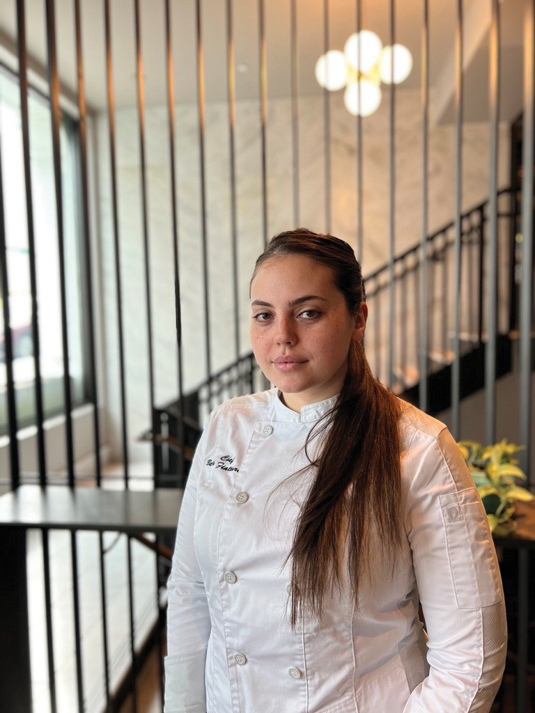 A woman in a white chef coat stands in a modern, bright room with vertical railings and a staircase. Her hair is tied back, and she gazes calmly at the camera.