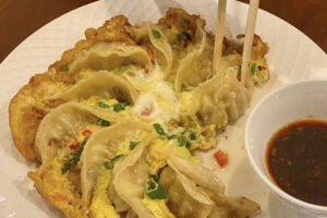 A plate of pan-fried dumplings arranged in a circle, cooked with egg and garnished with chopped vegetables, next to a small bowl of dipping sauce; chopsticks are picking up one dumpling.