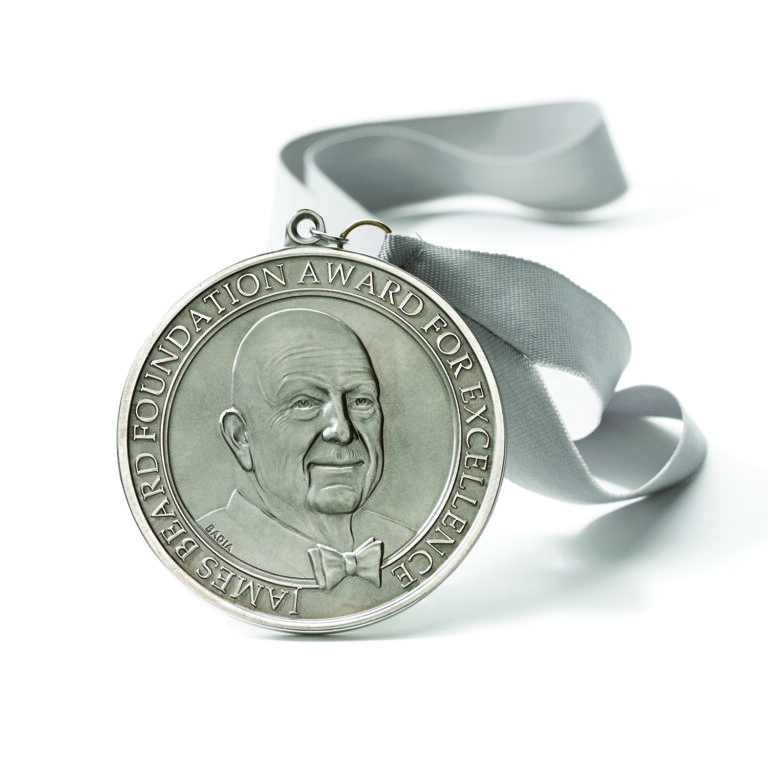 A silver medal with a gray ribbon, featuring a raised profile of an older man wearing a bow tie. The text around the edge reads, "James Beard Foundation Award for Excellence.