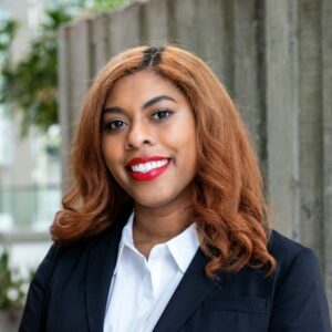 A woman with curly auburn hair and bright red lipstick is smiling. She is wearing a white shirt and a dark blazer, standing in front of a blurred outdoor background with greenery and concrete structures.